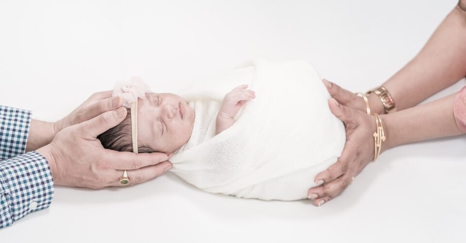 Panoramic photograph of both parents’ hands resting on either side of their newborn, asleep in white