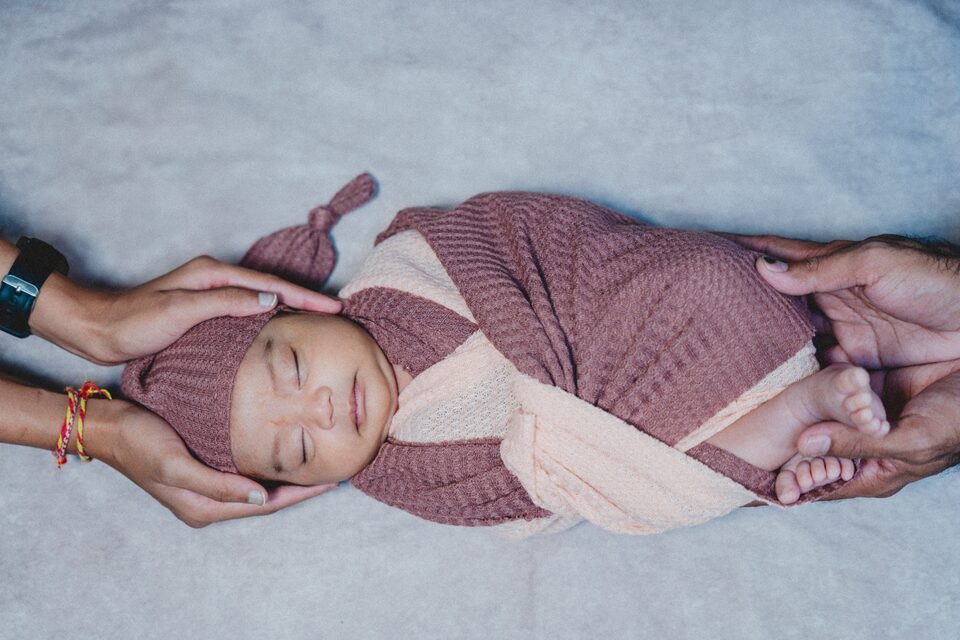 Newborn in a mauve wrap and knotted hat, supported on either side by a parent’s hands