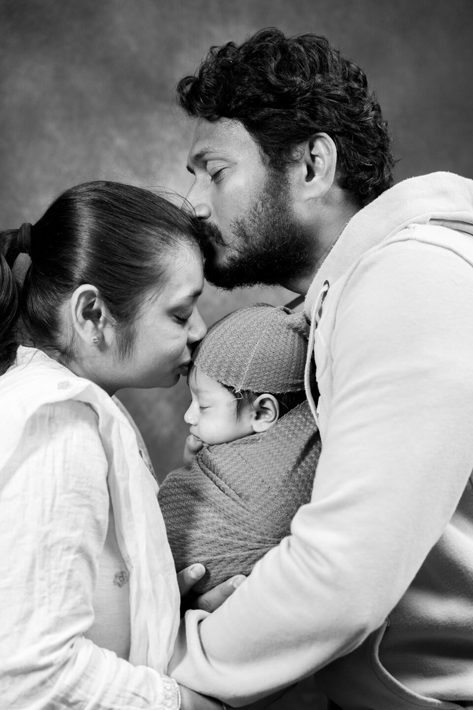 Black and white photograph of both parents leaning in to kiss their newborn at once