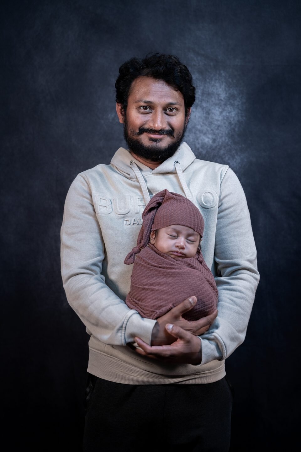 Father holding his newborn in a dusty rose wrap, lit softly against a dark backdrop