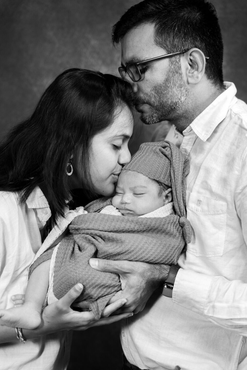 Black and white photograph of a father kissing the top of the mother’s head while she holds their newborn