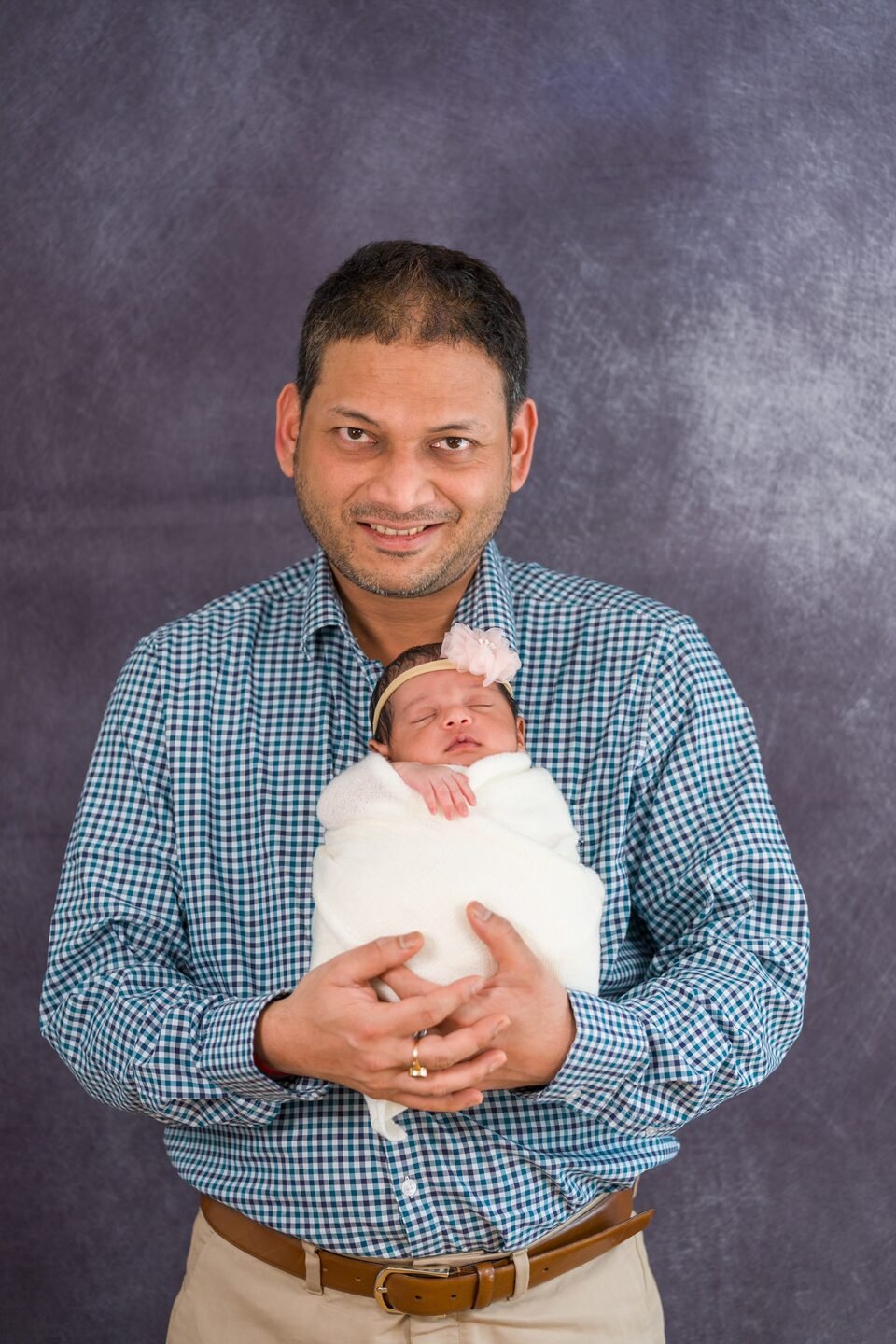 Father cradling his newborn, wrapped in white, against a deep studio backdrop