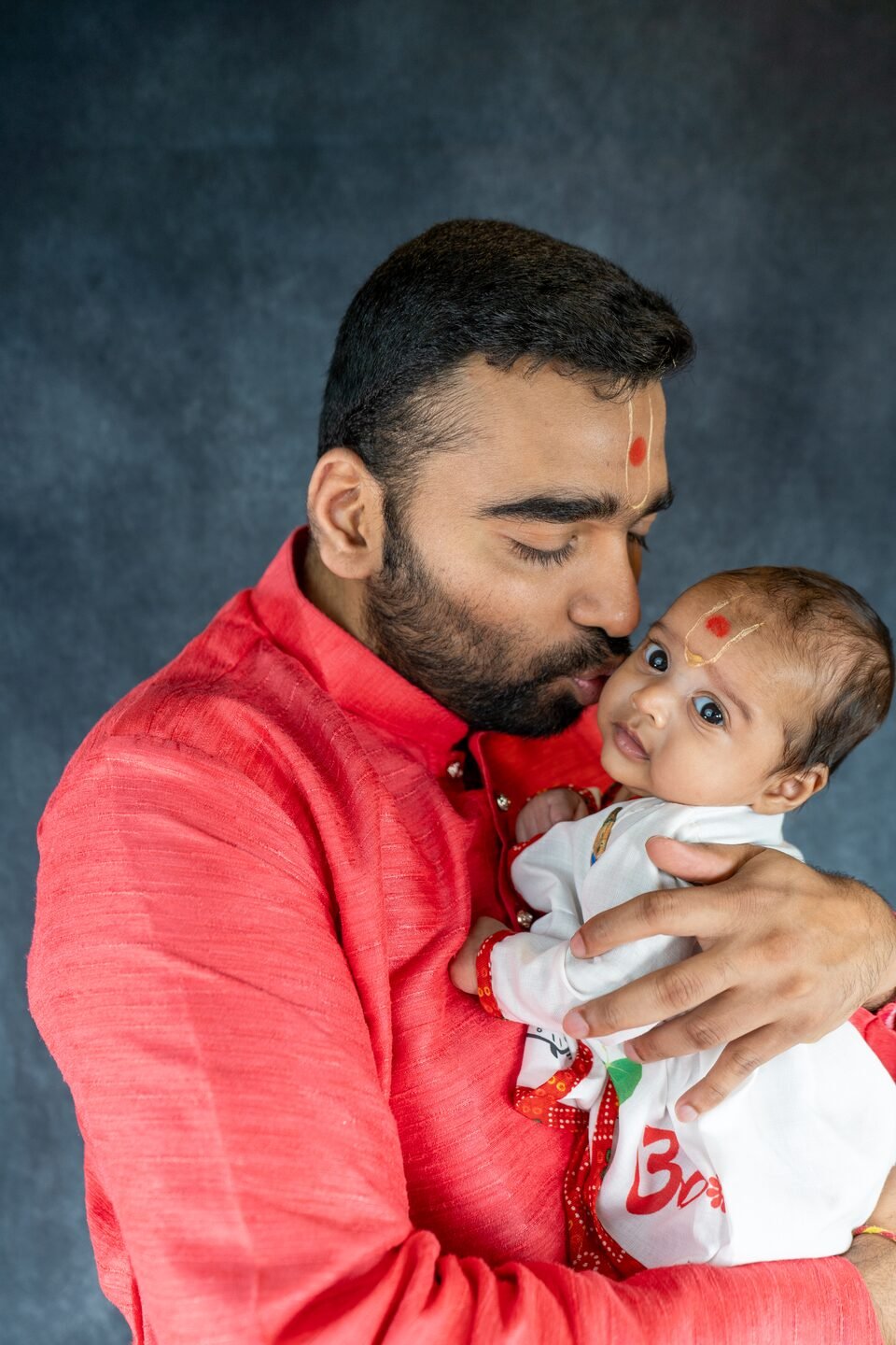 Father in a red kurta kissing his newborn on the cheek