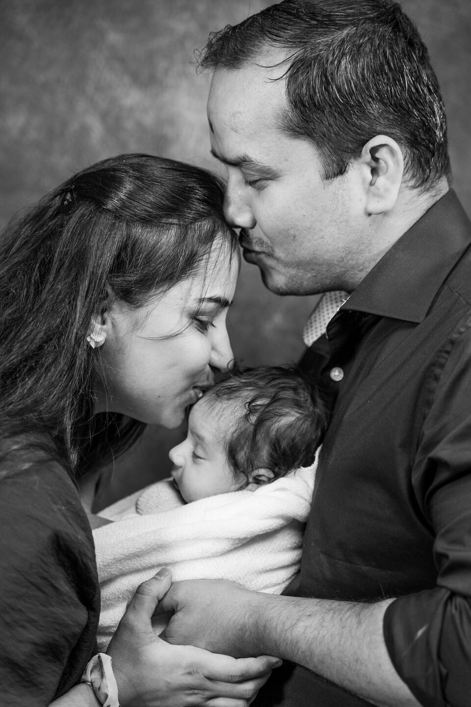 Black and white photograph of a father kissing the mother on the forehead as she holds their newborn