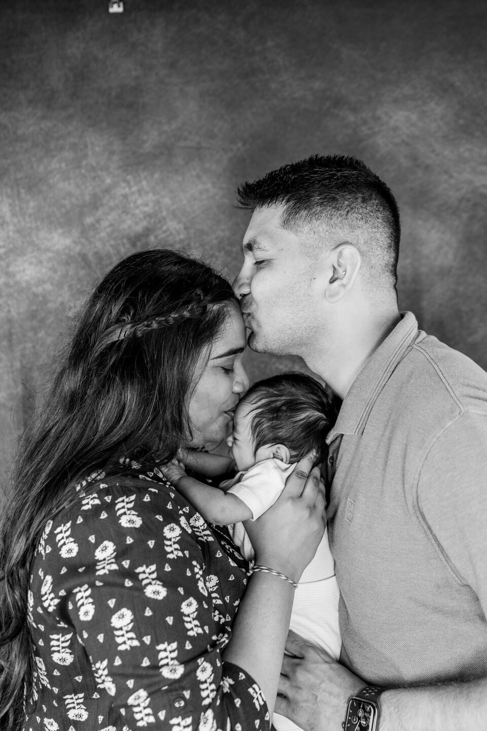 Black and white portrait of parents leaning towards each other with their baby held between them