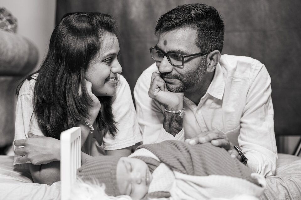 Black and white photograph of two parents lying down and looking at each other, their newborn asleep in front of them
