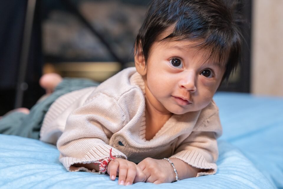 Baby in a pale pink cardigan propped on a blue blanket, looking straight at the camera