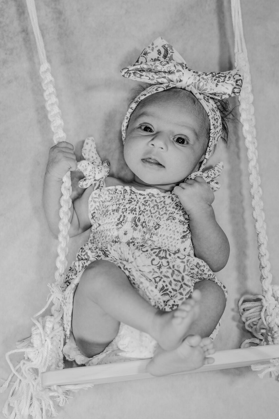 Black and white photograph of a baby girl in a lace romper and bow headband, seated on a wooden swing