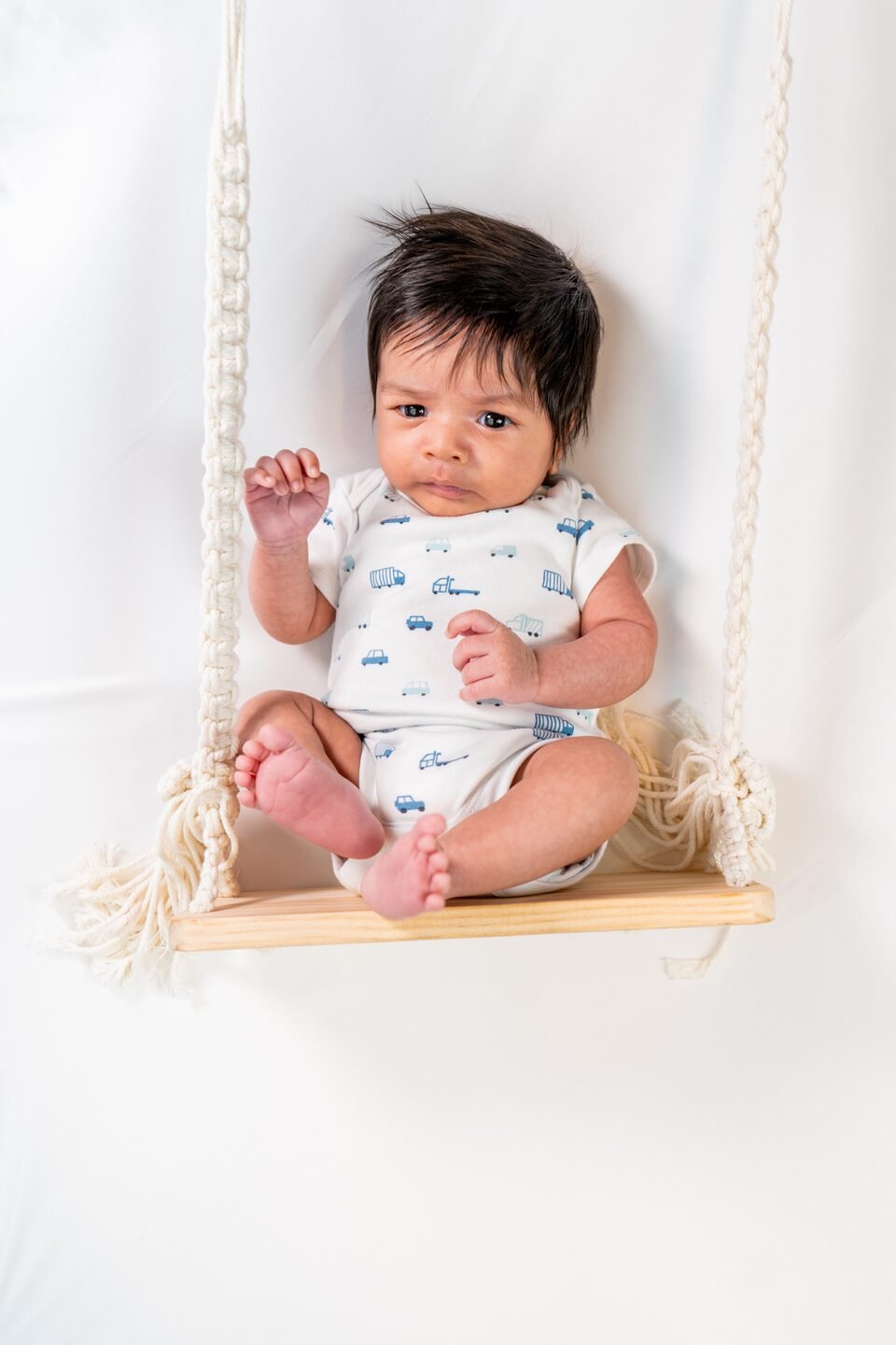 Baby in a white patterned onesie sitting upright on a wooden swing