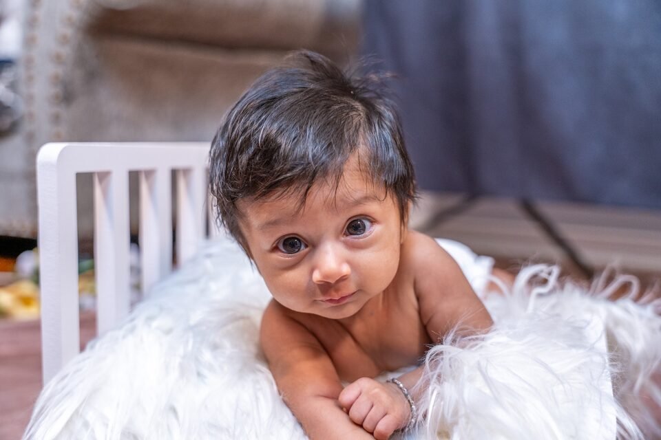 Baby lying on white fur inside a small wooden crib, head lifted towards the camera
