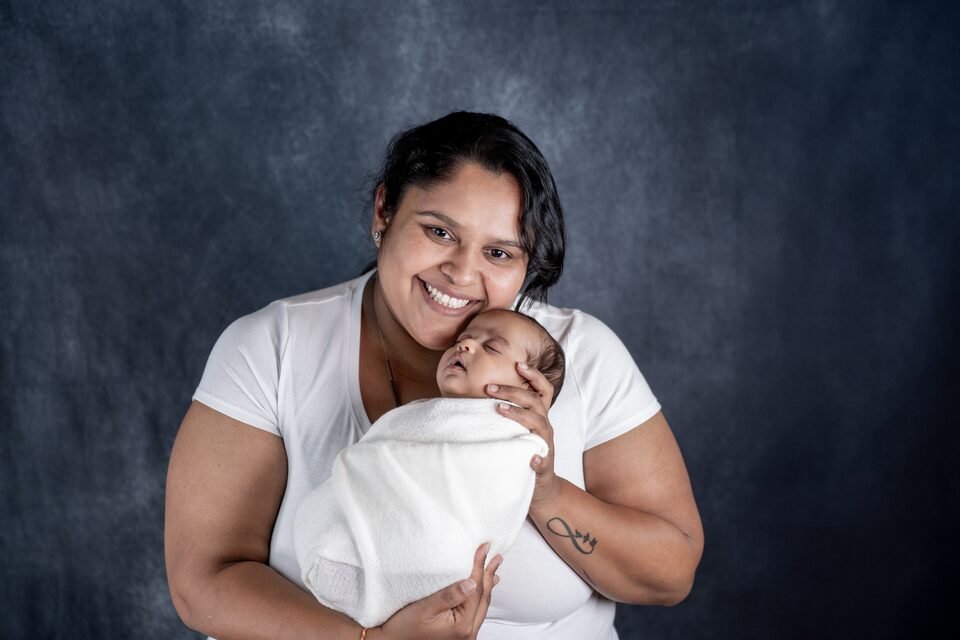 Mother smiling as she holds her newborn, wrapped in white, against a blue studio backdrop
