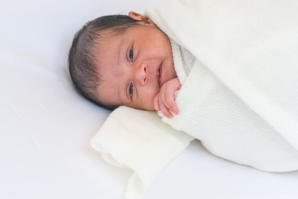 Wide-awake newborn resting a hand against her cheek, wrapped in soft cream fabric