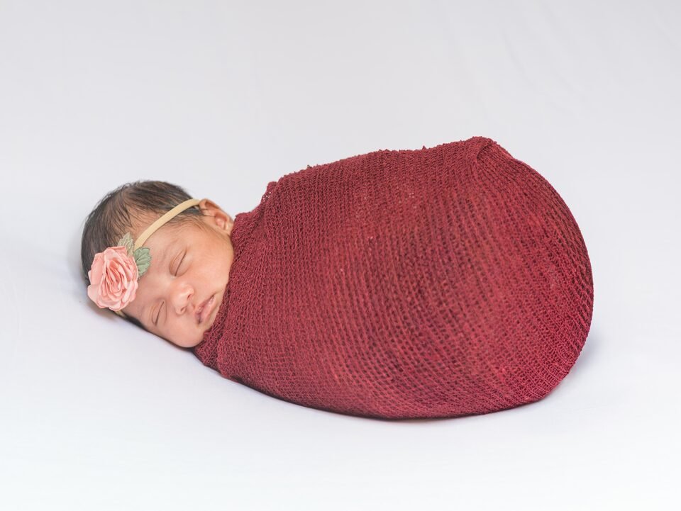Newborn wrapped in deep burgundy with a small floral headband, asleep