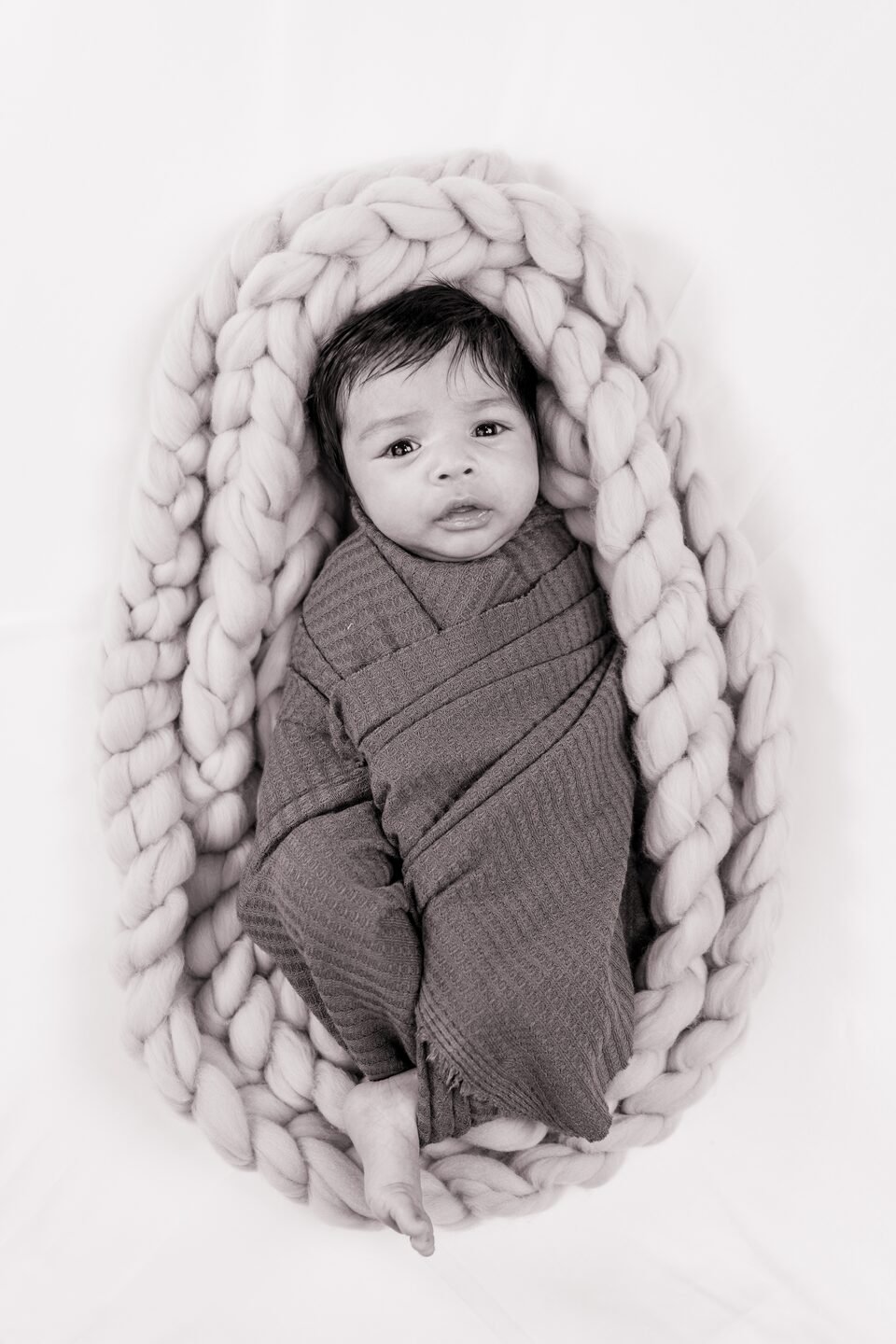 Newborn in a charcoal wrap resting inside a pale grey braided wool nest