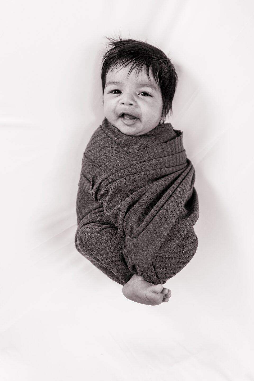 Newborn swaddled in a charcoal ribbed wrap on white bedding, eyes closed