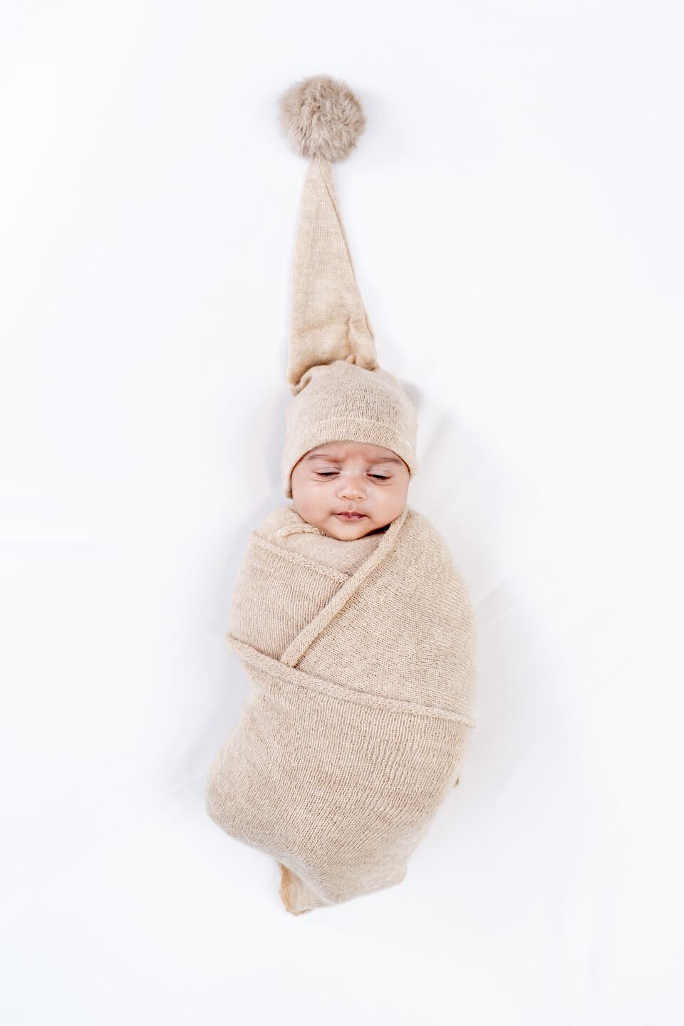 Sleeping newborn swaddled in a cream knit wrap with a long pompom hat, on a pale studio backdrop