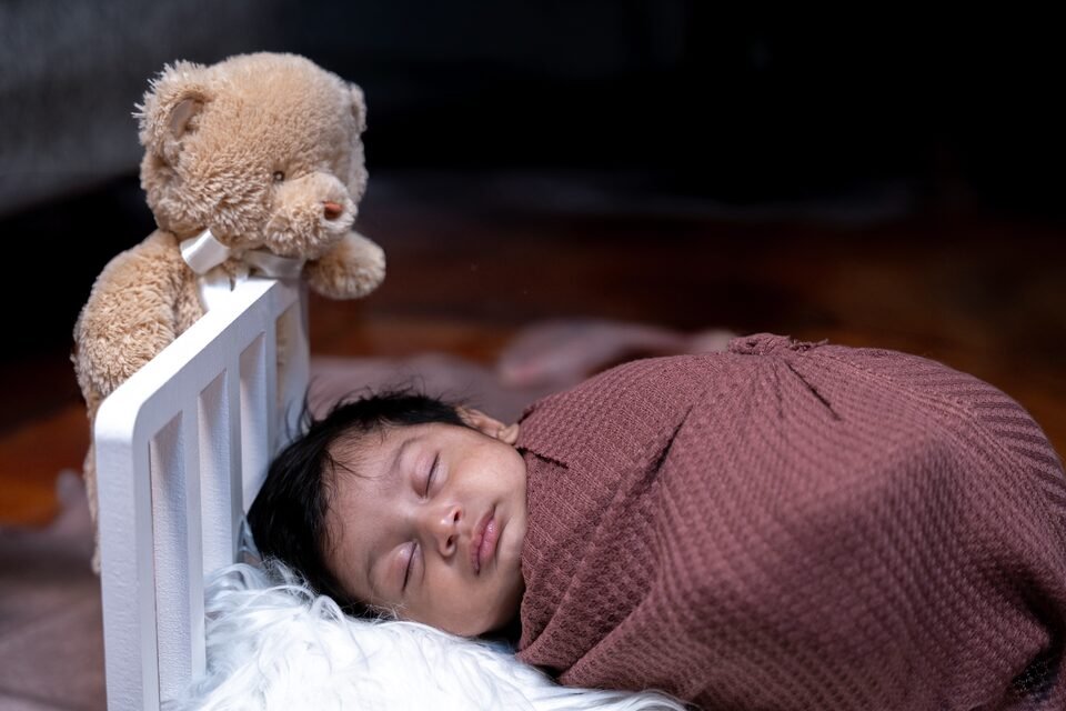 Newborn asleep in a small wooden crib on white fur, a knitted teddy resting at the head of the bed