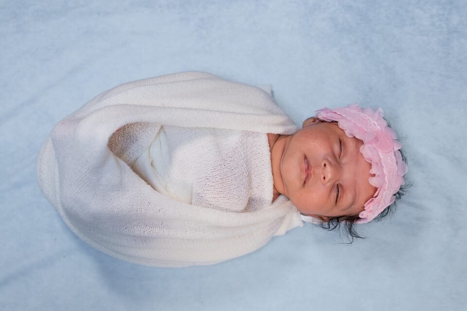 Newborn swaddled in cream with a pale pink floral headband, on a dusty blue backdrop