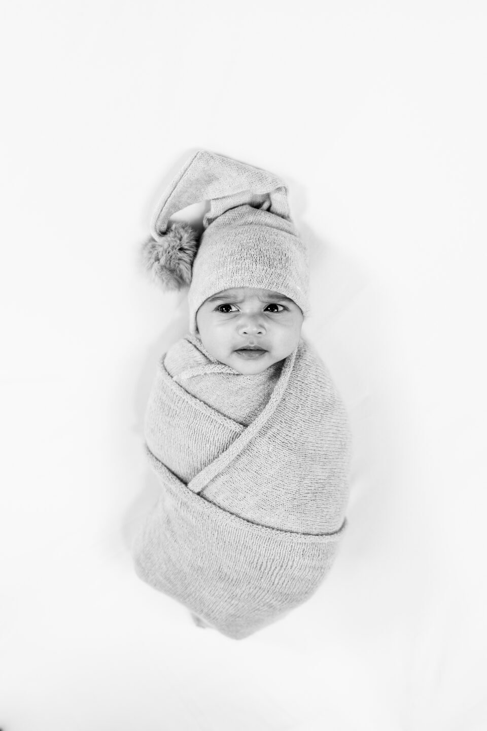 Black and white portrait of a newborn swaddled in grey with a soft knotted hat, eyes open