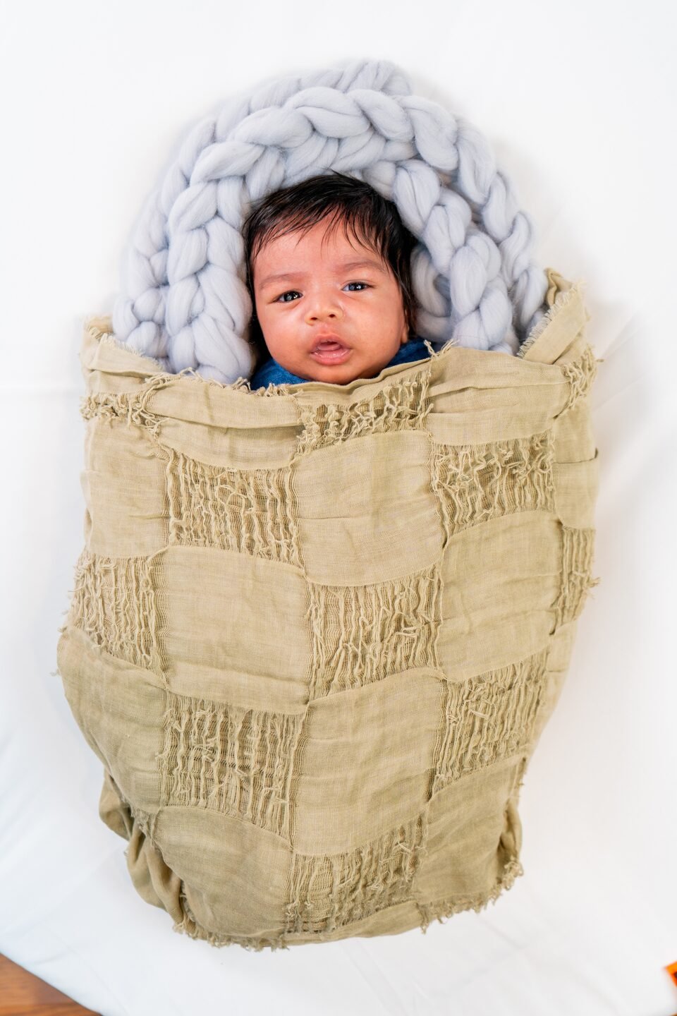 Newborn wrapped in textured khaki linen inside a soft grey wool nest