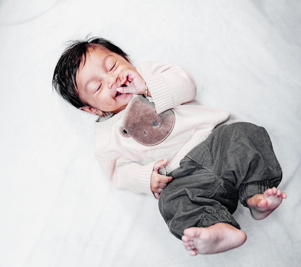 Newborn in a cream knit top and olive trousers, asleep with a pacifier on white bedding