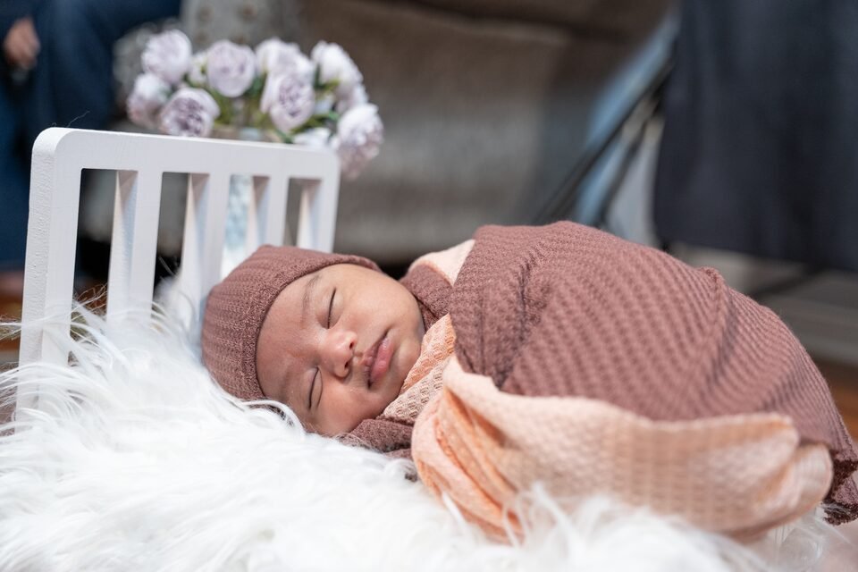 Newborn in a mauve knit wrap asleep on white fur in a small white bed, pale flowers behind