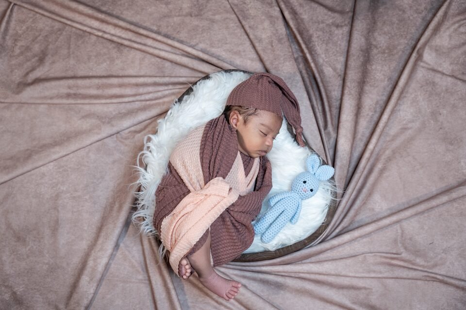 Newborn in a mauve wrap asleep beside a small blue knitted bunny