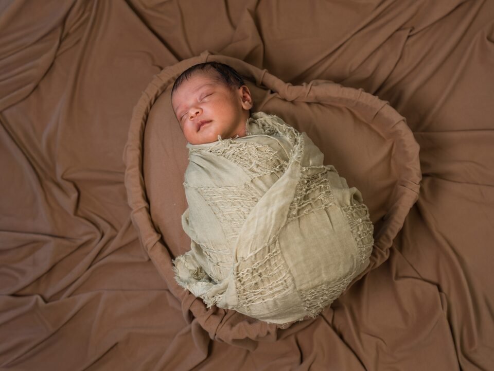 Newborn in an olive linen wrap settled into a fabric nest on a warm brown backdrop