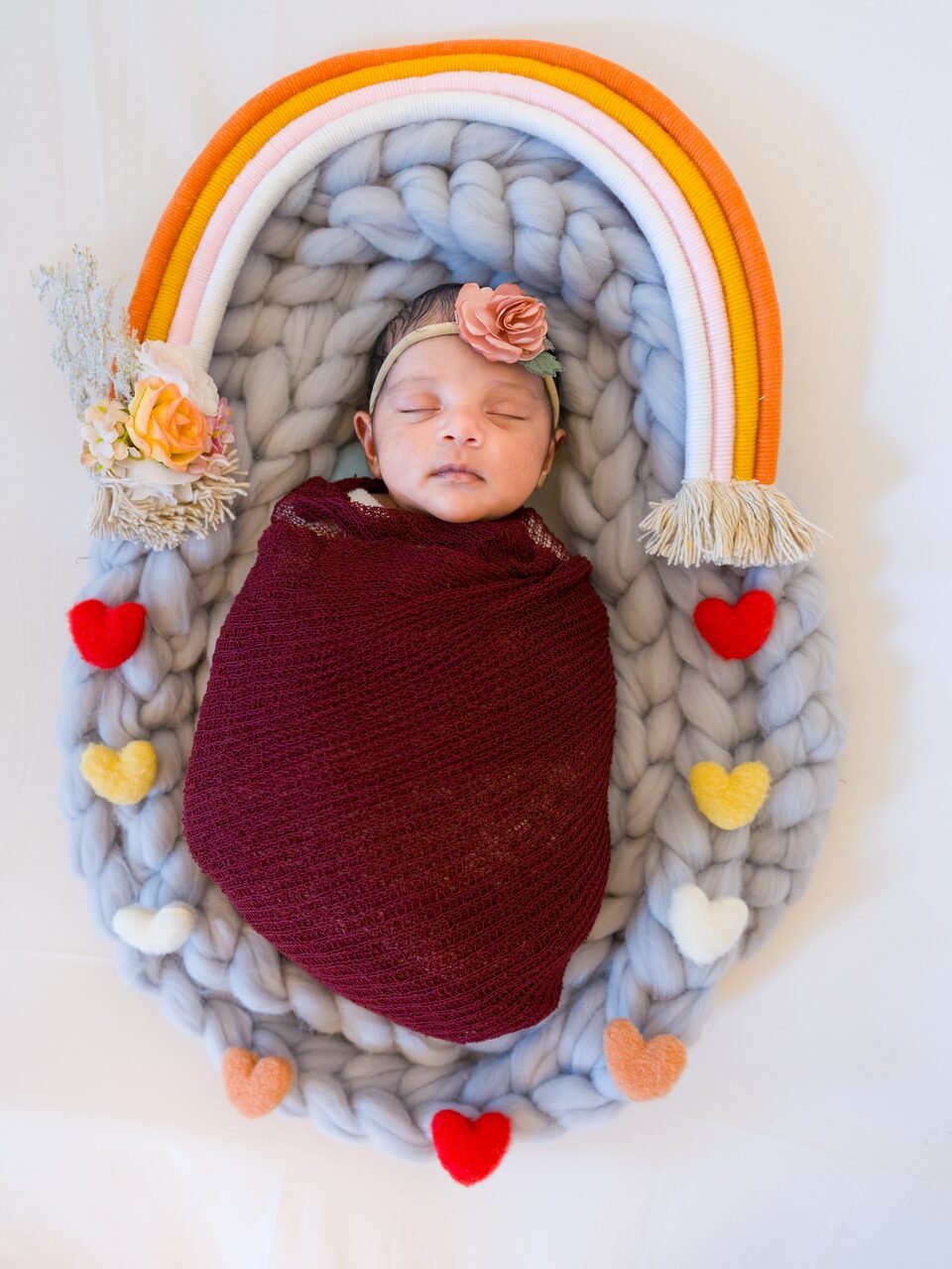 Newborn photographed from above, wrapped in burgundy inside a braided wool nest ringed with a soft rainbow of felt