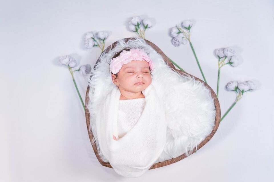 Newborn asleep in a shallow wooden bowl lined with white fur, framed by pale blossom stems
